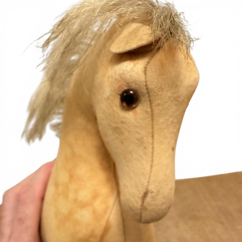 May include: A stuffed horse toy made of light tan fabric with visible stitched seams along the face and neck. The horse features a textured, light-colored synthetic mane and dark brown, round, glossy plastic eyes set into the fabric head.