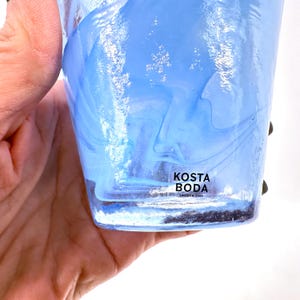 Puede incluir: Un vaso de vidrio transparente con un dise&ntilde;o en espiral azul claro y blanco. El vaso tiene una etiqueta que dice "KOSTA BODA SWEDEN 1742". El vaso est&aacute; sostenido por una mano.