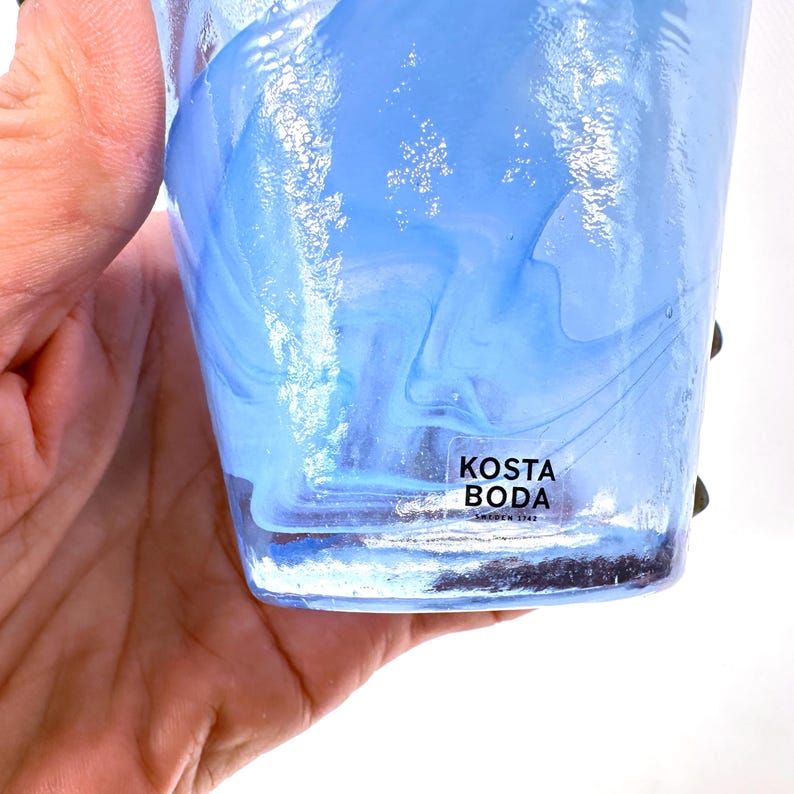 Puede incluir: Un vaso de vidrio transparente con un dise&ntilde;o en espiral azul claro y blanco. El vaso tiene una etiqueta que dice "KOSTA BODA SWEDEN 1742". El vaso est&aacute; sostenido por una mano.
