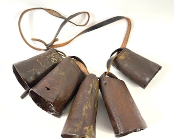 May include: A set of four weathered, dark brown metal cowbells suspended from a thin, worn brown leather strap. Each bell has a cylindrical, slightly tapered shape with visible rust, metallic texture, and a small clapper hanging from the center.