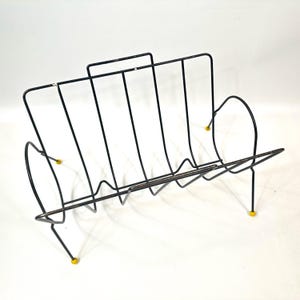 May include: Black metal magazine rack with a mid-century modern design. The rack features vertical bars and curved sides, with yellow feet. The rack is designed to hold magazines, books, or other reading materials.