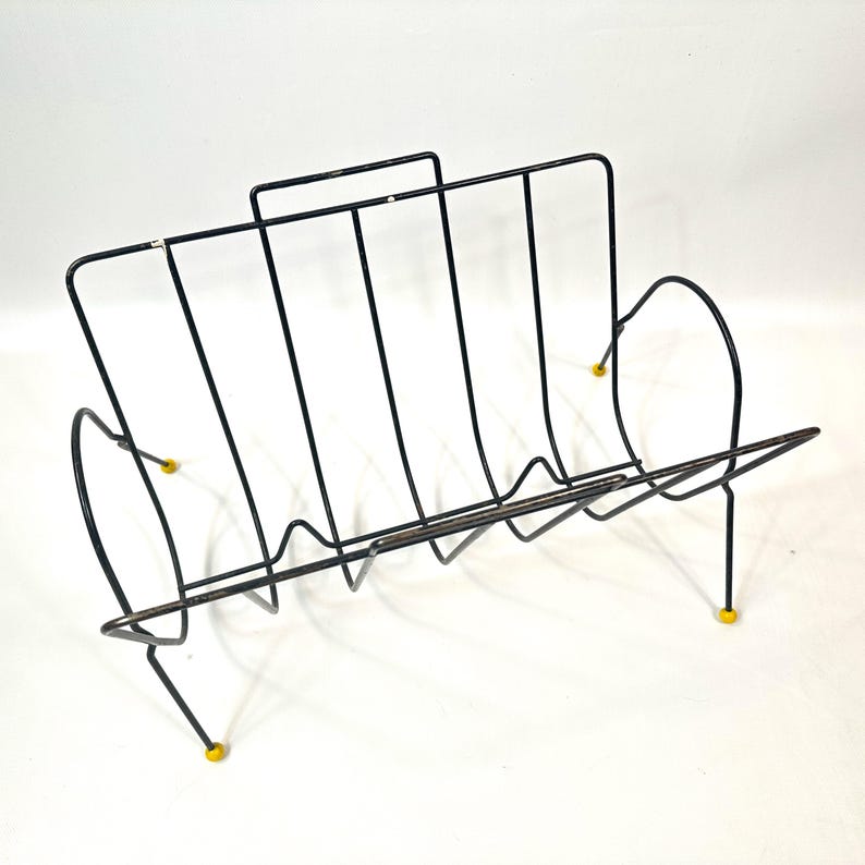 May include: Black metal magazine rack with a mid-century modern design. The rack features vertical bars and curved sides, with yellow feet. The rack is designed to hold magazines, books, or other reading materials.