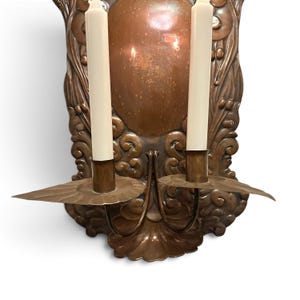 K&ouml;nnte beinhalten: Ein Wandkerzenhalter aus Kupfer mit einer ovalen Mittelplatte, die mit gepr&auml;gten Schn&ouml;rkeln und floralen Motiven verziert ist. Zwei geschwungene Arme ragen aus der Basis hervor, die jeweils eine runde Tropfschale und eine hohe, cremewei&szlig;e Kerze tragen.