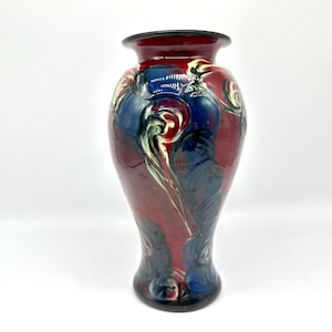 May include: A decorative ceramic vase with a flared rim and base. The vase features a maroon background with blue, white, and black abstract designs. The vase is approximately 10 inches tall.