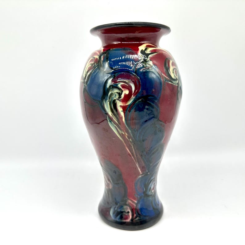 May include: A decorative ceramic vase with a flared rim and base. The vase features a maroon background with blue, white, and black abstract designs. The vase is approximately 10 inches tall.