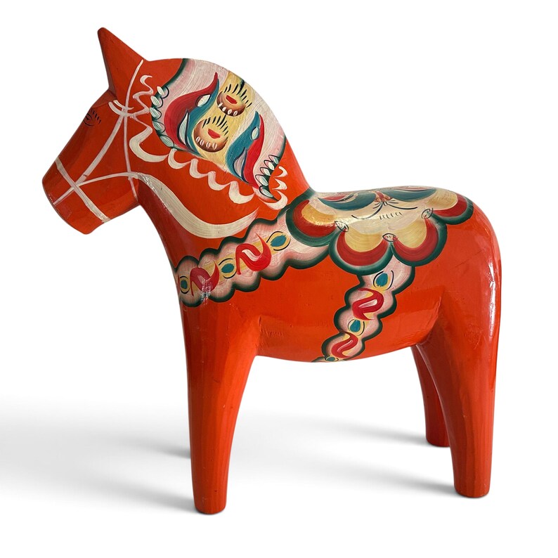 Puede incluir: Una figura de caballo de madera de color naranja brillante con detalles pintados a mano en blanco, verde azulado, verde y rojo. El dise&ntilde;o incluye una brida pintada, un patr&oacute;n de silla de montar decorativo en el lomo y motivos florales estilizados.