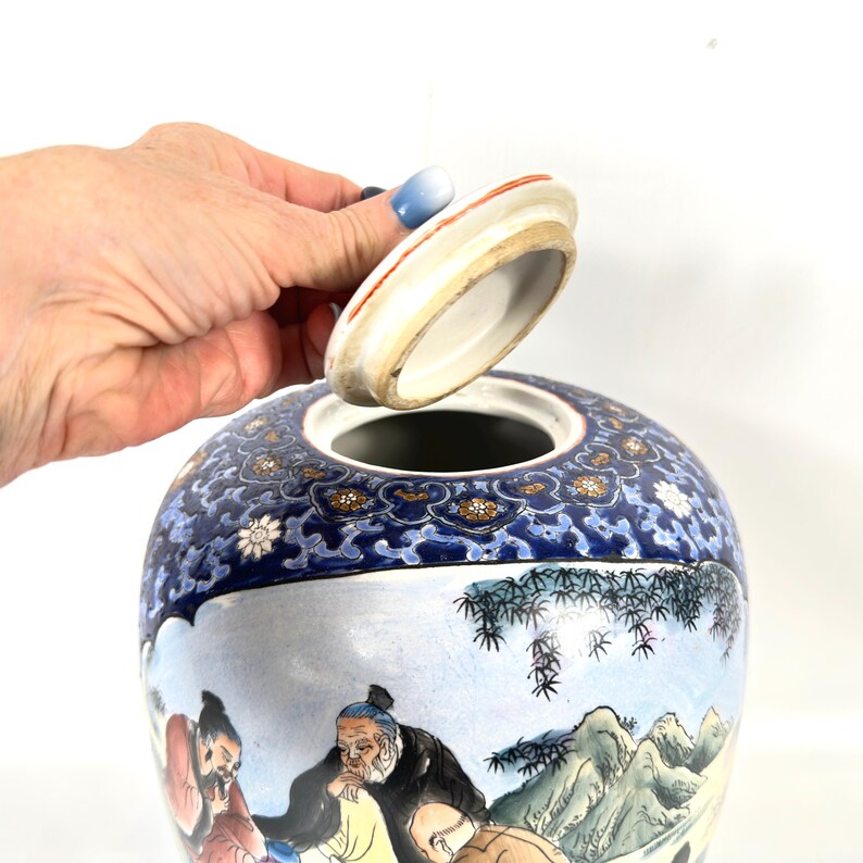 Puede incluir: Un tarro de jengibre de cer&aacute;mica con tapa extra&iacute;ble, que presenta una escena pintada a mano de figuras en un paisaje con monta&ntilde;as y bamb&uacute;. La parte superior tiene un borde decorativo azul oscuro con patrones florales en oro, blanco y azul.