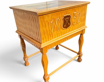 May include: A light brown wooden chest with a textured, carved exterior. The front features the carved numbers 19 and 52 flanking a central shield emblem with crossed arrows. The chest stands on four carved legs with a connecting base frame.