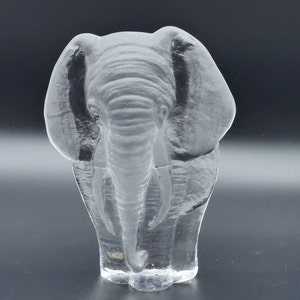 May include: A clear glass elephant figurine with detailed trunk, tusks, and ears. The sculpture is transparent, allowing light to pass through, and is set against a dark background. The elephant is a decorative item.