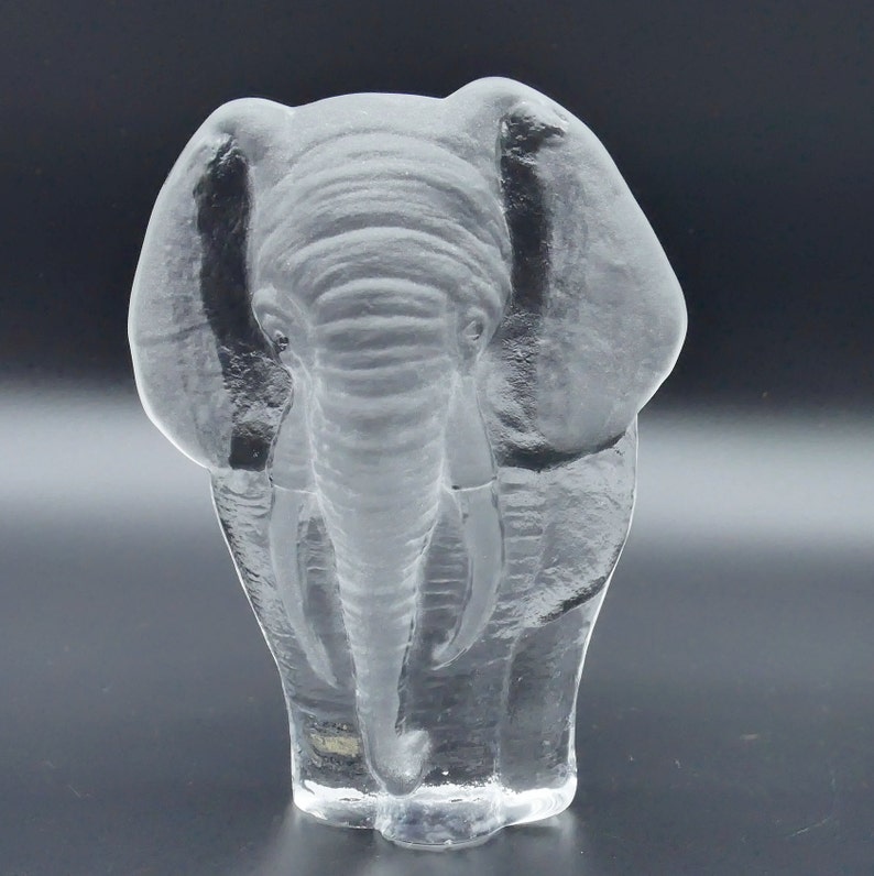 May include: A clear glass elephant figurine with detailed trunk, tusks, and ears. The sculpture is transparent, allowing light to pass through, and is set against a dark background. The elephant is a decorative item.