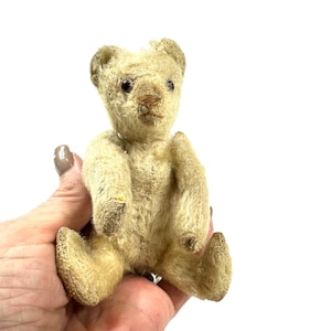 May include: A small, vintage-style teddy bear with worn, light beige fur. The bear features dark, round eyes, a stitched nose, and rounded ears. Its limbs are articulated, and the fur appears textured and aged, showing signs of wear on the paws.