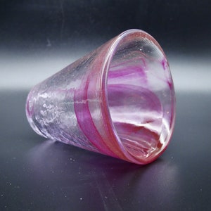 Puede incluir: Un vaso de vidrio transparente con forma acampanada, con bandas arremolinadas de color magenta y rosa. El vidrio tiene un aspecto texturizado y descansa sobre una superficie oscura. El dise&ntilde;o le da al vaso un aspecto art&iacute;stico &uacute;nico.