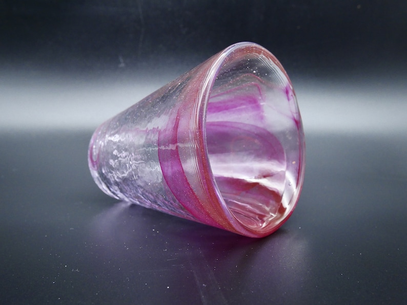 Puede incluir: Un vaso de vidrio transparente con forma acampanada, con bandas arremolinadas de color magenta y rosa. El vidrio tiene un aspecto texturizado y descansa sobre una superficie oscura. El dise&ntilde;o le da al vaso un aspecto art&iacute;stico &uacute;nico.