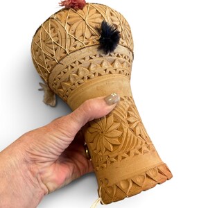 May include: A tan clay drum featuring intricate hand-carved geometric patterns, including star-like floral motifs and triangular borders. The drum head is secured with light-coloured string and decorated with small, fuzzy pink and black pom-poms.