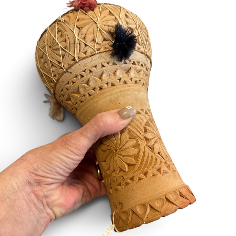 May include: A tan clay drum featuring intricate hand-carved geometric patterns, including star-like floral motifs and triangular borders. The drum head is secured with light-coloured string and decorated with small, fuzzy pink and black pom-poms.