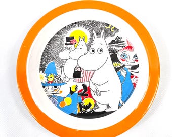 May include: A round, white plate with an orange rim featuring a Moomin illustration. The illustration depicts several white, cartoon-like characters with a black and white background, including a yellow sun and black trees.