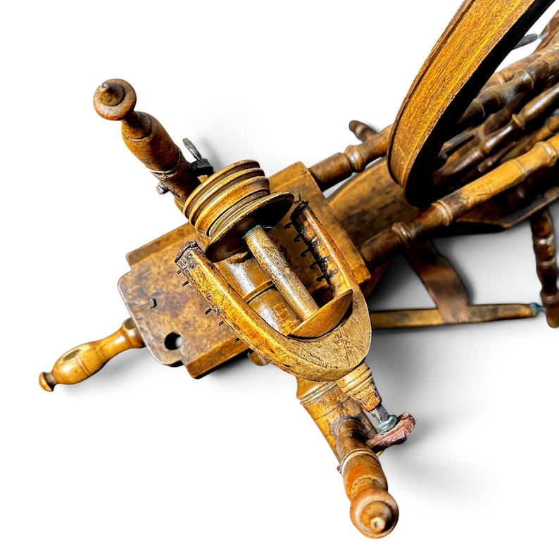May include: A wooden spinning wheel featuring a central flyer assembly with a bobbin and whorl, connected to a large drive wheel. The structure is crafted from light brown wood with turned details, including vertical spindles and a horizontal base.