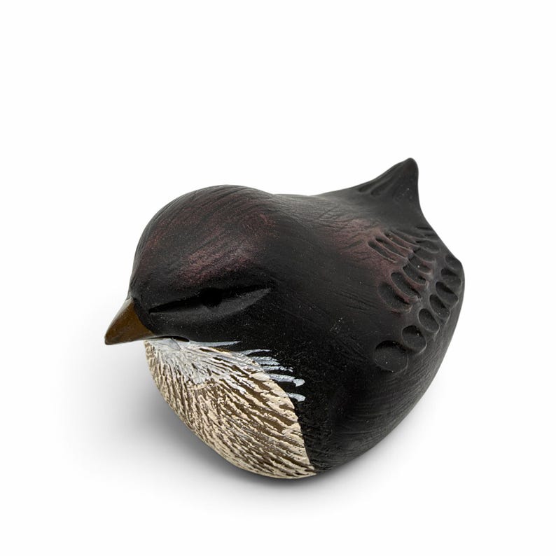 May include: A small, hand-carved wooden bird figurine with a dark brown, almost black finish. The bird features a pointed beak, carved wing details, and a textured, light-colored chest area with visible white paint strokes on the front.
