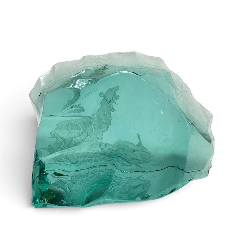 Peut inclure: Une sculpture en verre bleu sarcelle translucide en forme de roche dentel&eacute;e, pr&eacute;sentant un motif grav&eacute; d'un loup hurlant debout sur une surface rocheuse. Le verre poss&egrave;de des bords irr&eacute;guliers et facett&eacute;s ainsi qu'une surface avant lisse.