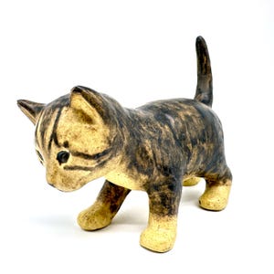 Op de afbeelding: Een keramieken beeldje van een tabby-katje met een lichtbruine basis en donkerbruine strepen. Het katje staat in een loopstand met zijn staart omhoog. De ogen zijn zwart en de oren zijn puntig.