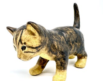 May include: A ceramic tabby kitten figurine with a light tan base and dark brown stripes. The kitten is depicted walking with its tail raised. The eyes are black, and the ears are pointed.