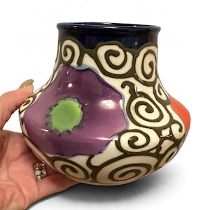 Puede incluir: Un jarrón de cerámica con base ancha y cuello estrecho, con un borde azul oscuro. El cuerpo está decorado con una flor morada con centro verde claro, rodeada de patrones de espirales marrón oscuro sobre un fondo color crema.