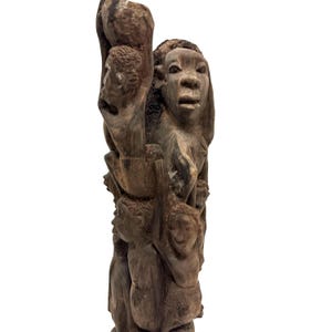 May include: A tall, dark brown wooden sculpture featuring a vertical arrangement of multiple human figures carved in relief. The figures are stacked and intertwined, displaying various facial expressions and postures within the textured wood grain.