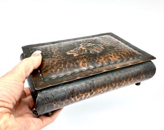 May include: A hammered copper box with a hinged lid. The lid features a raised design of a moose. The box has a dark patina and is rectangular with rounded edges. The box is held in a hand.