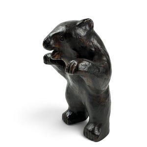 May include: A dark brown bear figurine standing on its hind legs with its front paws raised. The sculpture features a textured, glossy surface with visible carved details on the face, ears, and paws, capturing a dynamic, upright posture.