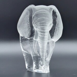 May include: A clear glass elephant sculpture with detailed trunk, tusks, and ears. The sculpture is a transparent, three-dimensional representation of an elephant, showcasing the animal's features. The base has text that reads "KOSTA BODA SWEDEN".