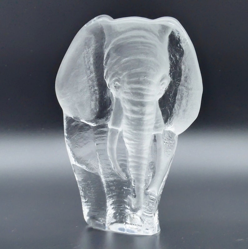 May include: A clear glass elephant sculpture with detailed trunk, tusks, and ears. The sculpture is a transparent, three-dimensional representation of an elephant, showcasing the animal's features. The base has text that reads "KOSTA BODA SWEDEN".