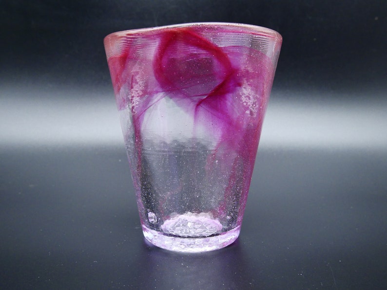 Puede incluir: Un vaso de vidrio transparente con forma acampanada, con dise&ntilde;os internos magenta y blancos arremolinados. La base y el borde tienen un aspecto texturizado. El vaso mide unos 10 cm de alto.