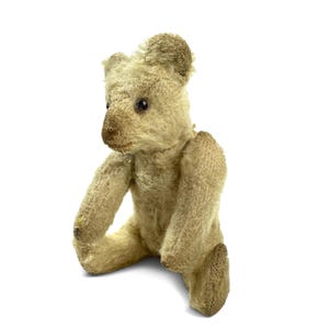May include: A vintage-style teddy bear crafted from light beige, textured mohair-like fabric. The bear features dark, round eyes, a stitched nose, and articulated limbs. It is shown in a seated position with a worn, weathered appearance.