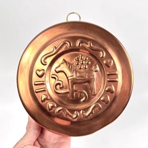 Peut inclure: Une décoration murale ronde de couleur cuivrée avec un motif de cheval au centre. Le cheval est entouré d'éléments décoratifs, notamment des cœurs et des motifs floraux. Une petite boucle dorée est en haut pour l'accrocher.