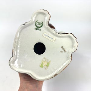 Peut inclure: Vue du dessous d'une figurine en c&eacute;ramique vintage. La base est blanche avec une bordure marron et un grand trou noir. La figurine porte une &eacute;tiquette indiquant "GOTEBORG" et "PRICE KENSINGTON MADE IN ENGLAND".