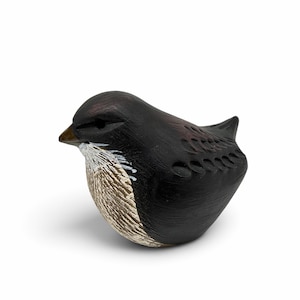 May include: A small, hand-carved wooden bird figurine featuring a dark brown and black finish. The bird has a textured, light brown chest with white accents, a pointed beak, and carved wing details along its back, presented in a side profile.