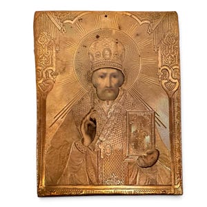 May include: A rectangular gold-toned metal icon featuring a central embossed figure of a bearded saint wearing a mitre and ornate vestments. The figure holds a book and makes a gesture of blessing, surrounded by a radiant halo and intricate border.