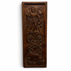 May include: A rectangular dark brown wooden wall panel featuring a hand-carved relief. The top section depicts a standing figure holding a staff and a horn, while the lower section shows a goat amidst stylized foliage and circular motifs.