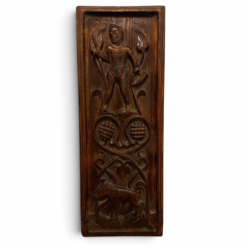 May include: A rectangular dark brown wooden wall panel featuring a hand-carved relief. The top section depicts a standing figure holding a staff and a horn, while the lower section shows a goat amidst stylized foliage and circular motifs.