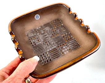 May include: A square brown ceramic tray with a wavy edge and a central geometric pattern of raised dots and lines. A small circular sticker with the text Uppsala-Ekeby Sweden is visible on the surface of the tray, which has a smooth finish.