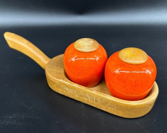 May include: A set of two orange ceramic salt and pepper shakers with wooden tops, sitting on a wooden tray with a handle.