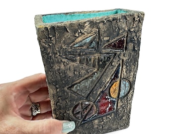 May include: A rectangular ceramic vase with a rough, dark brown textured exterior and a bright turquoise interior. The front surface features raised geometric shapes in shades of red, yellow, and blue, creating a tactile, abstract relief pattern.
