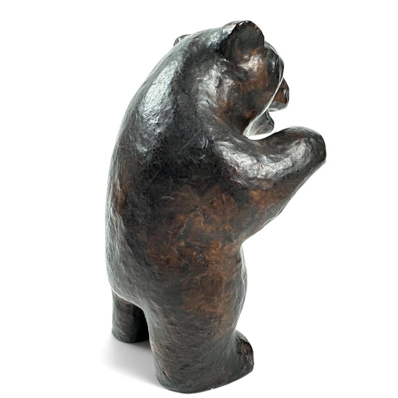 May include: A dark brown bear figurine standing on its hind legs with its front paws raised. The sculpture features a textured, uneven surface with a glossy finish, showing the bear in a profile view with its mouth slightly open and head turned.