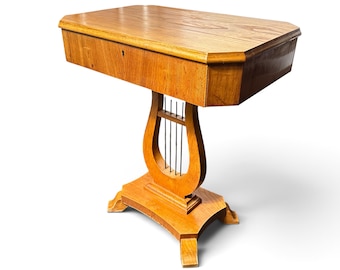 Peut inclure: Une table d'appoint en bois brun clair dotée d'un plateau rectangulaire aux coins coupés et d'un tiroir unique avec une serrure. La table repose sur un pied central en forme de lyre, agrémenté de fines tiges métalliques verticales.