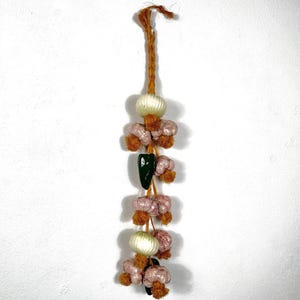 May include: A string of decorative, hand-crafted, fabric onions and peppers hanging from a brown twine. The onions are white and the peppers are pink and green.