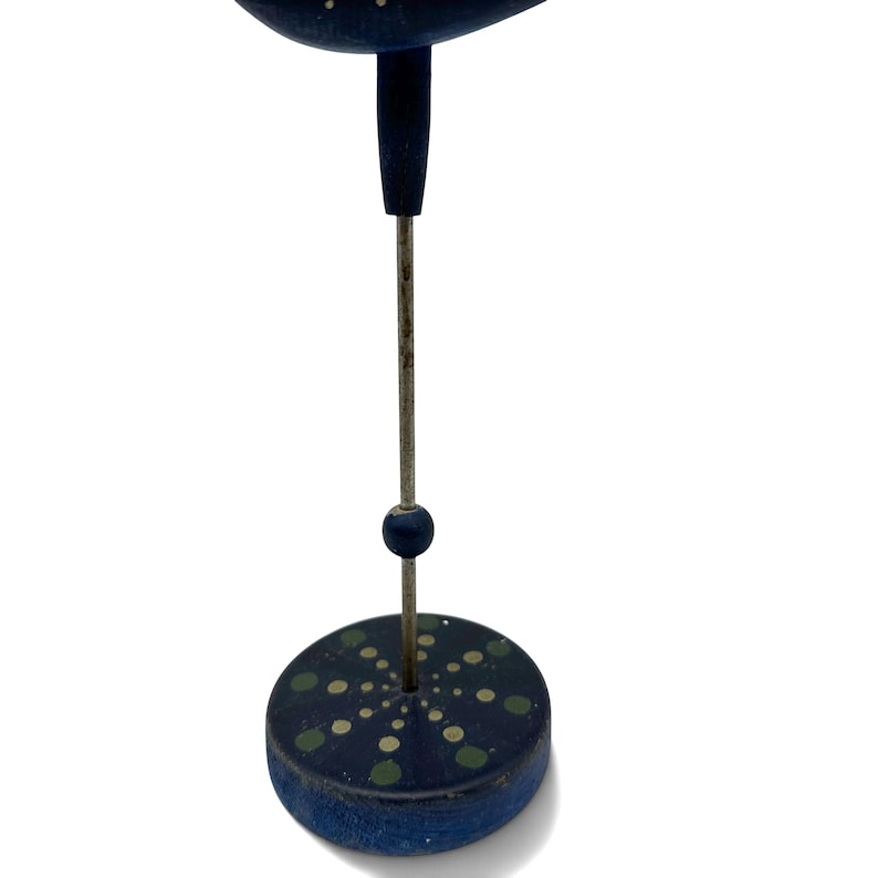 May include: A dark blue wooden bird figure mounted on a thin metal rod attached to a circular blue base. The base features a painted pattern of light green and white dots arranged in a radial design, with a small blue bead on the metal rod.