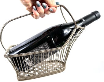 May include: Silver-coloured metal wine bottle holder with a handle, holding a dark wine bottle. The holder has a basket-like design with vertical bars and a decorative border. The bottle is angled within the holder.