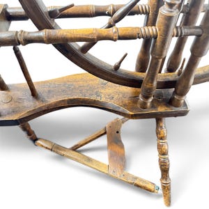 May include: A brown wooden spinning wheel featuring a large circular frame with spokes, a horizontal base with a foot treadle mechanism, and turned vertical supports. The wood displays a weathered finish with visible grain and structural joints.