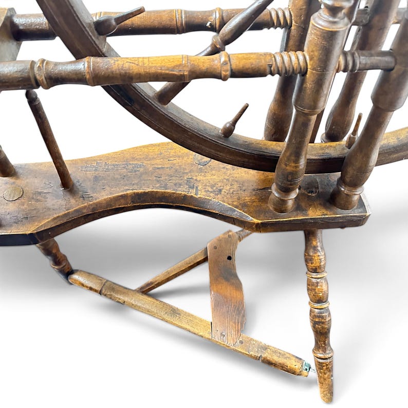 May include: A brown wooden spinning wheel featuring a large circular frame with spokes, a horizontal base with a foot treadle mechanism, and turned vertical supports. The wood displays a weathered finish with visible grain and structural joints.