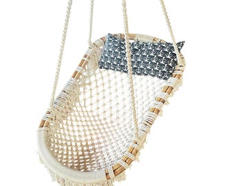 May include: A hanging macrame swing chair featuring a natural wood frame wrapped in cream-colored cord with a woven net seat. The chair includes a rectangular pillow with a repeating black and white cat face pattern and long fringe trim.